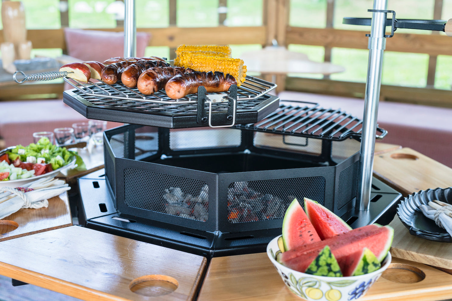 Sausage and corn sitting on the Rotating Grill accessory attached to the Kota Grill. Watermelon sits in a bowl on one of the tables attached to the grill.