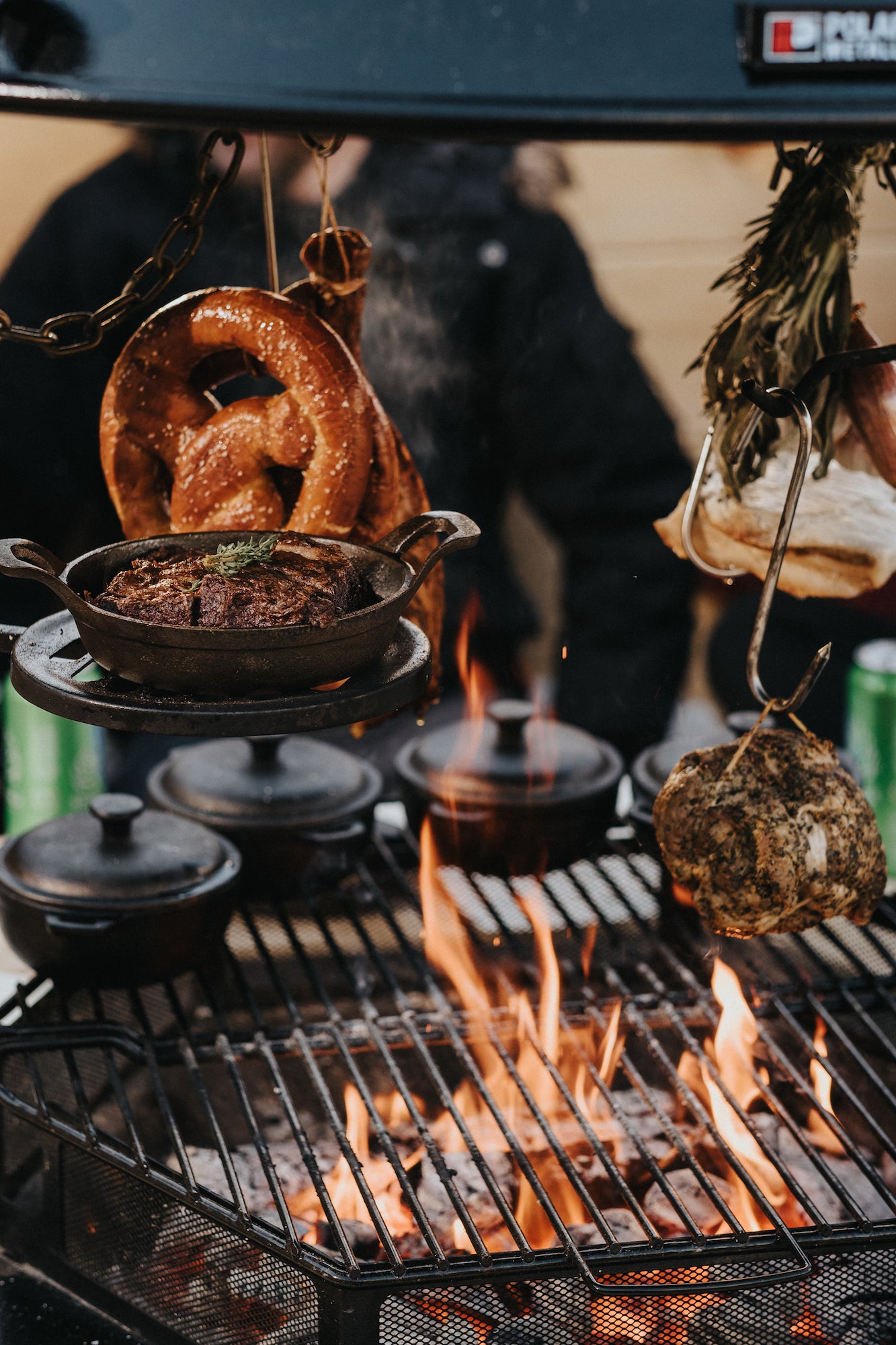      Pretzels hang off string with spices above a fire with braized pork waiting to be served. Naan bread hangs just above the fire.  