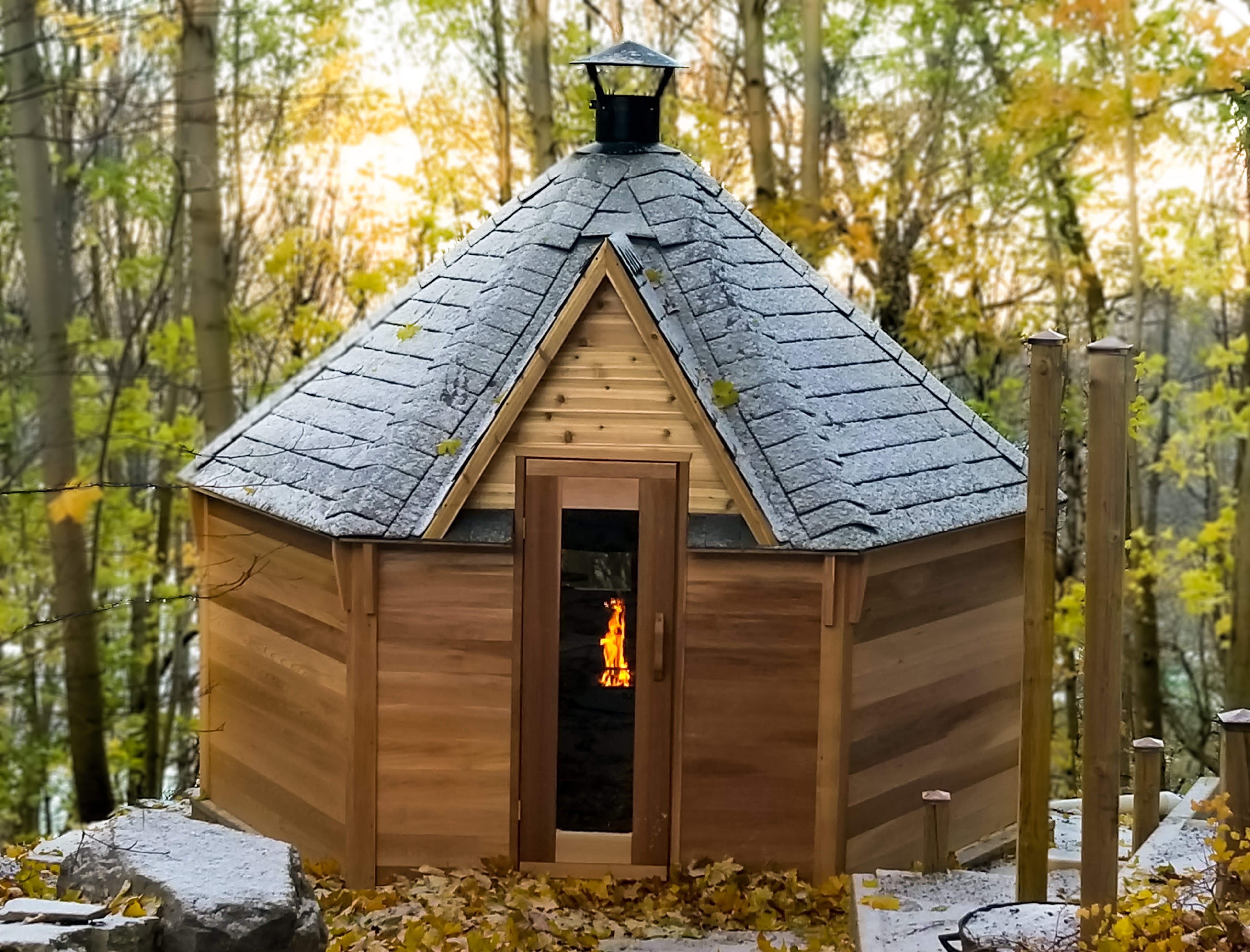 A red cedar Kota Cabin stands in a forested area with a fire burning visible through the pane glass door window.