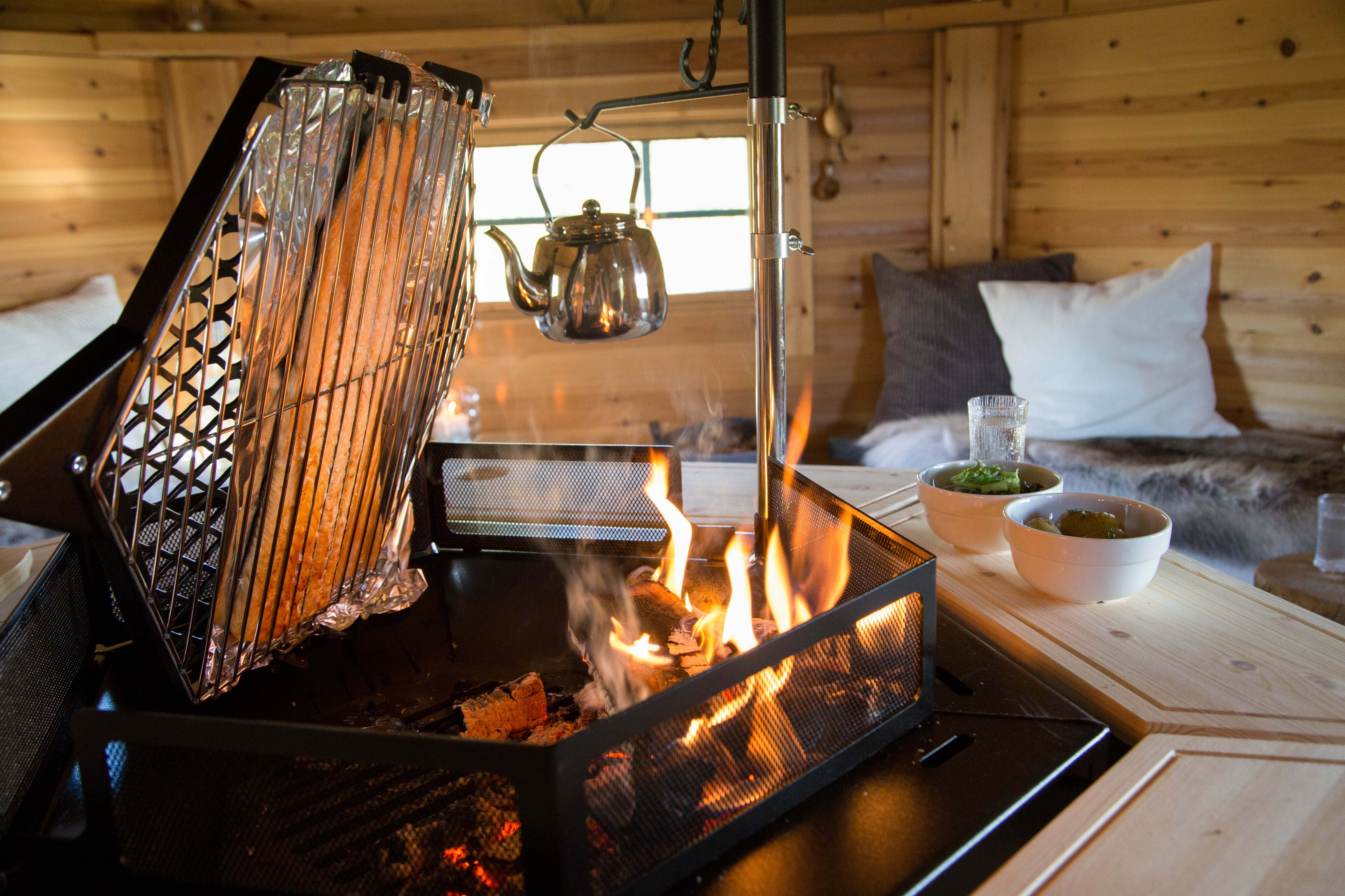 A close-up of a salmon cooking on the Rotating Grill Accessory, inside a hut. Garnishing for the food sits on the table, and pillows rest on the benches.