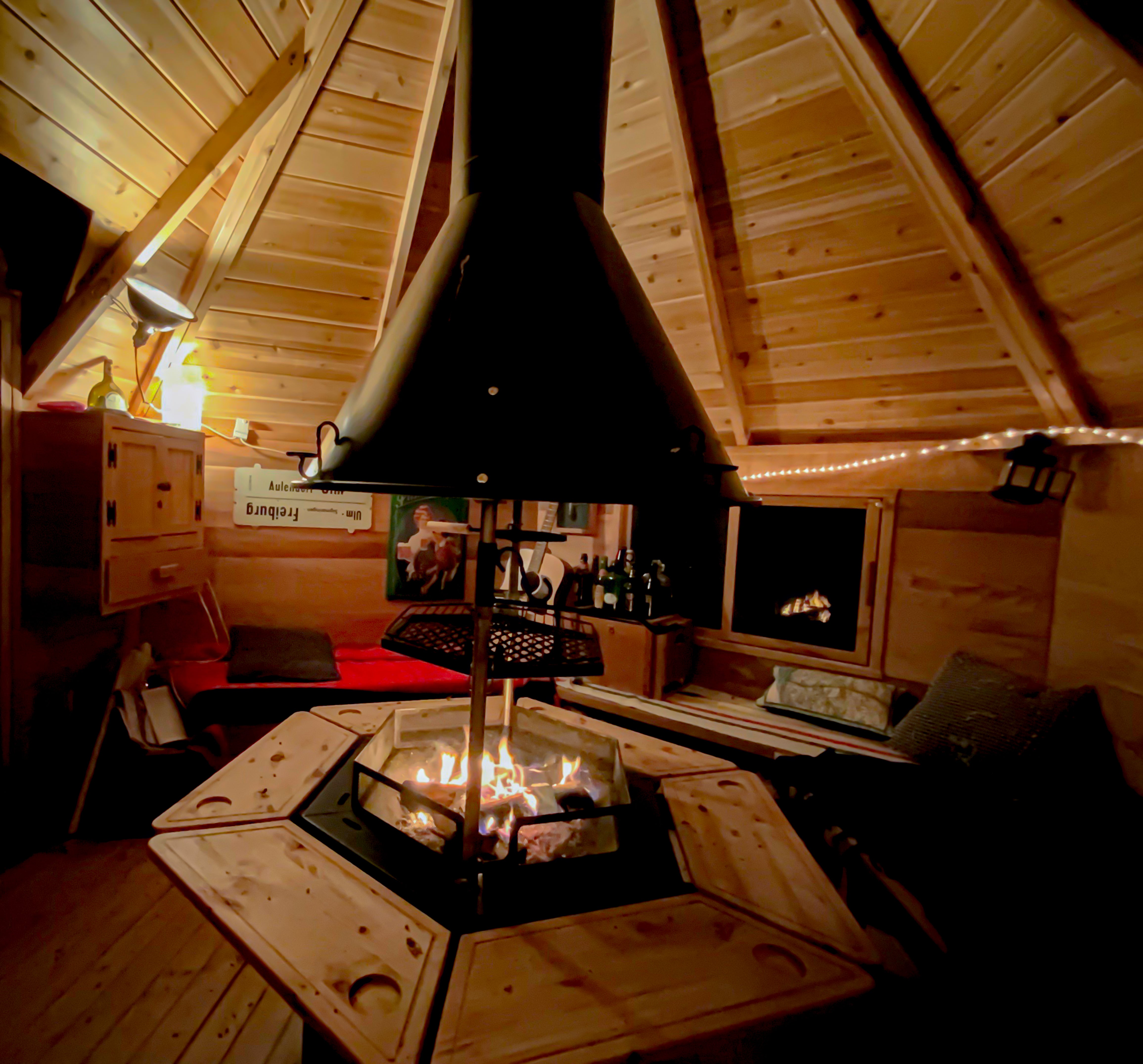     Fire and smoke rising through the chimney in the ceiling of the Cabin. Lights line the red cedar walls and pillows cover the wooden benches.   
