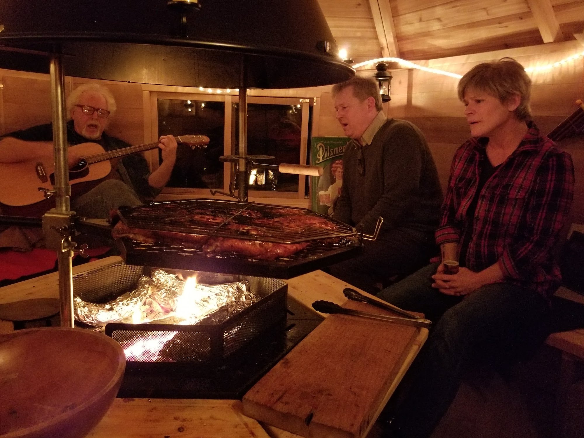   People singing and playing guitar in a Kota Cabin while assorted food roasts on the grill.     
