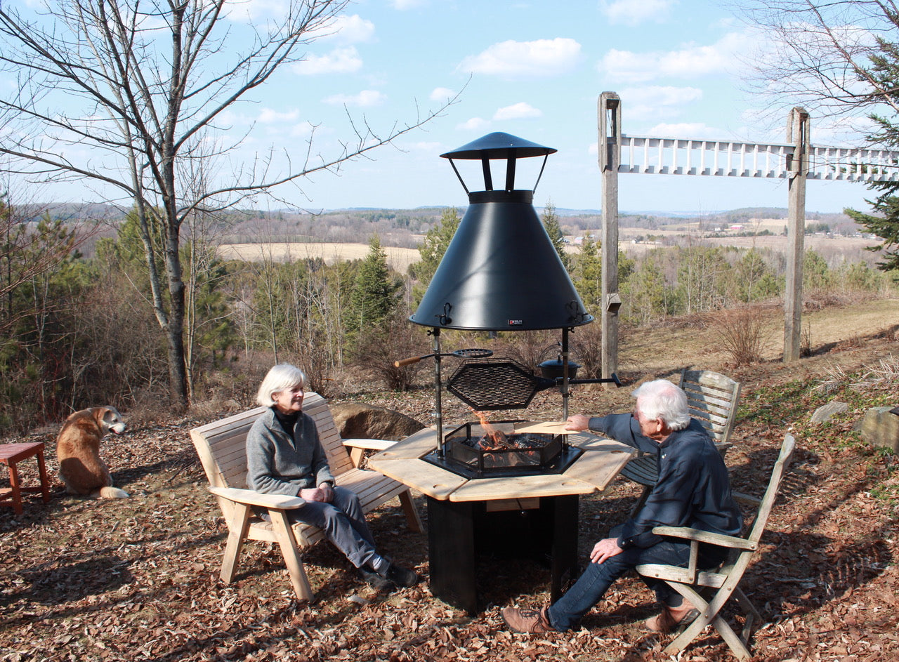 Two people sit around a Kota Grill fire in early spring while a dog sits overlooking a forest and fields.