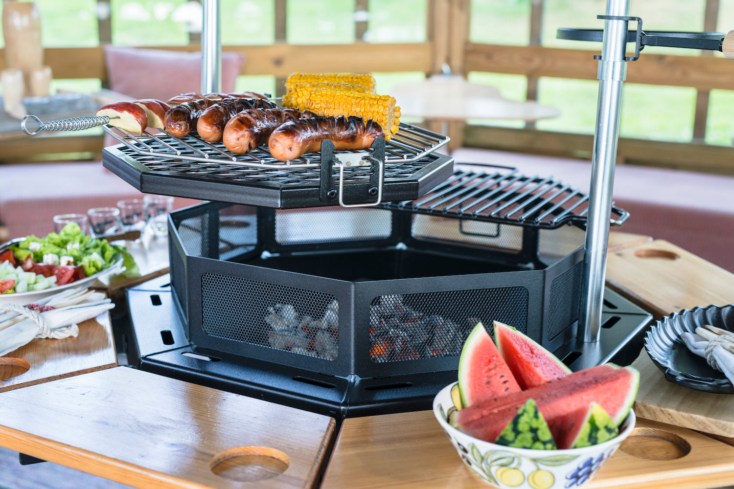    Sausage and corn sitting on the Rotating Grill accessory attached to the Kota Grill. Watermelon sits in a bowl on one of the tables attached to the grill.    
