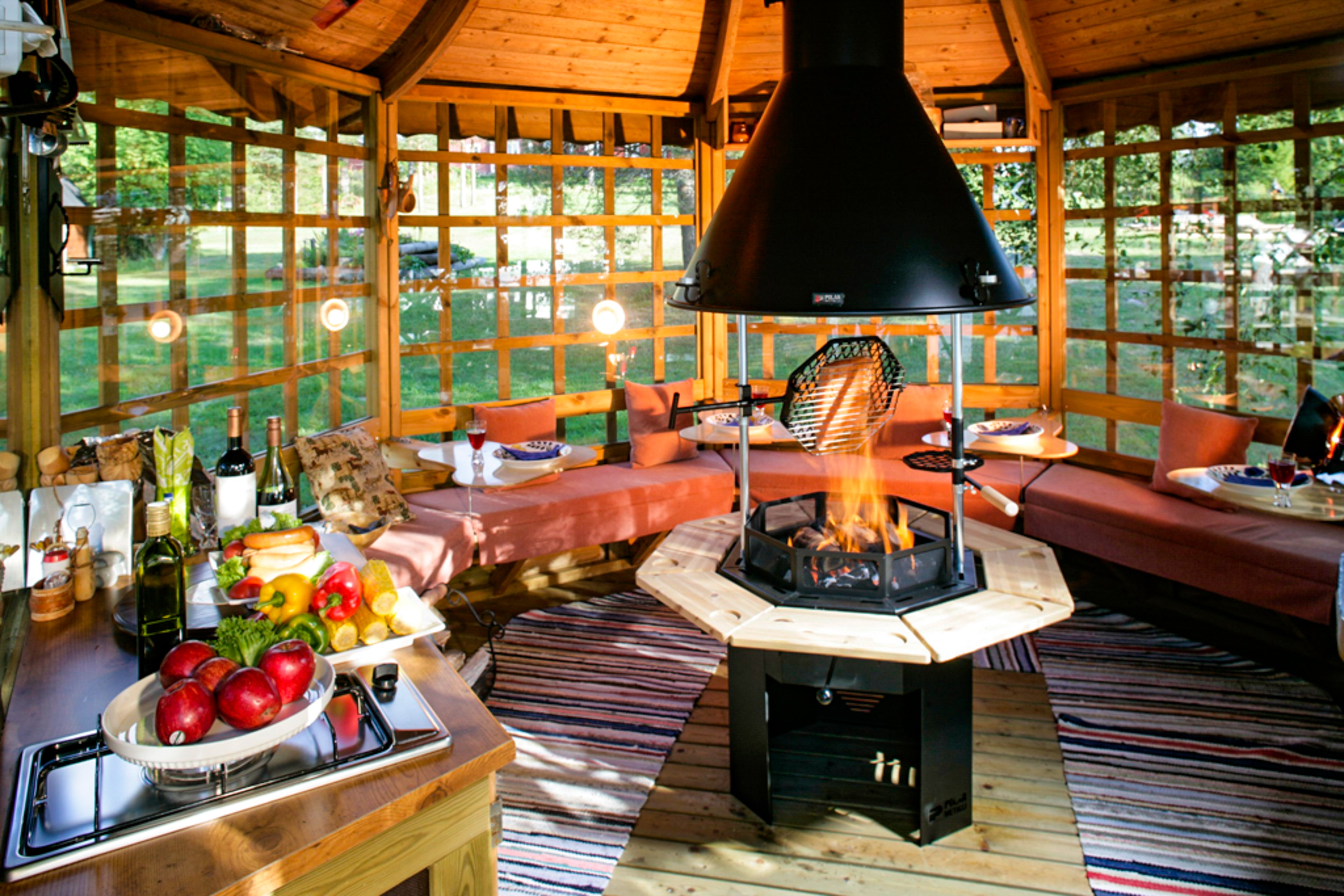   A Gazebo style structure encloses a series of pillowed seats and a Kota Grill. Food sits around the burning fire.     