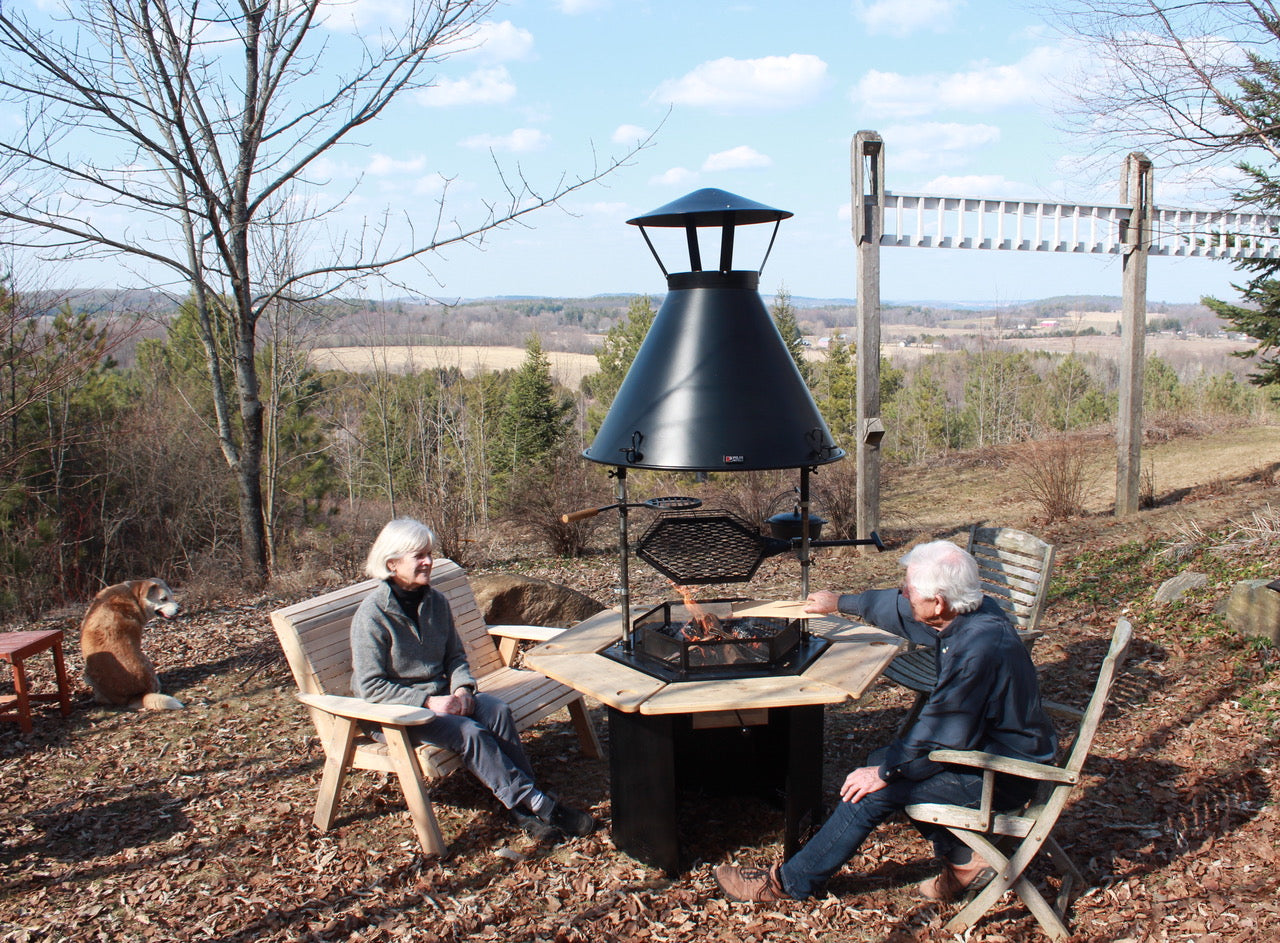    Two people sit around a Kota Grill fire in early spring while a dog sits overlooking a forest and fields.    