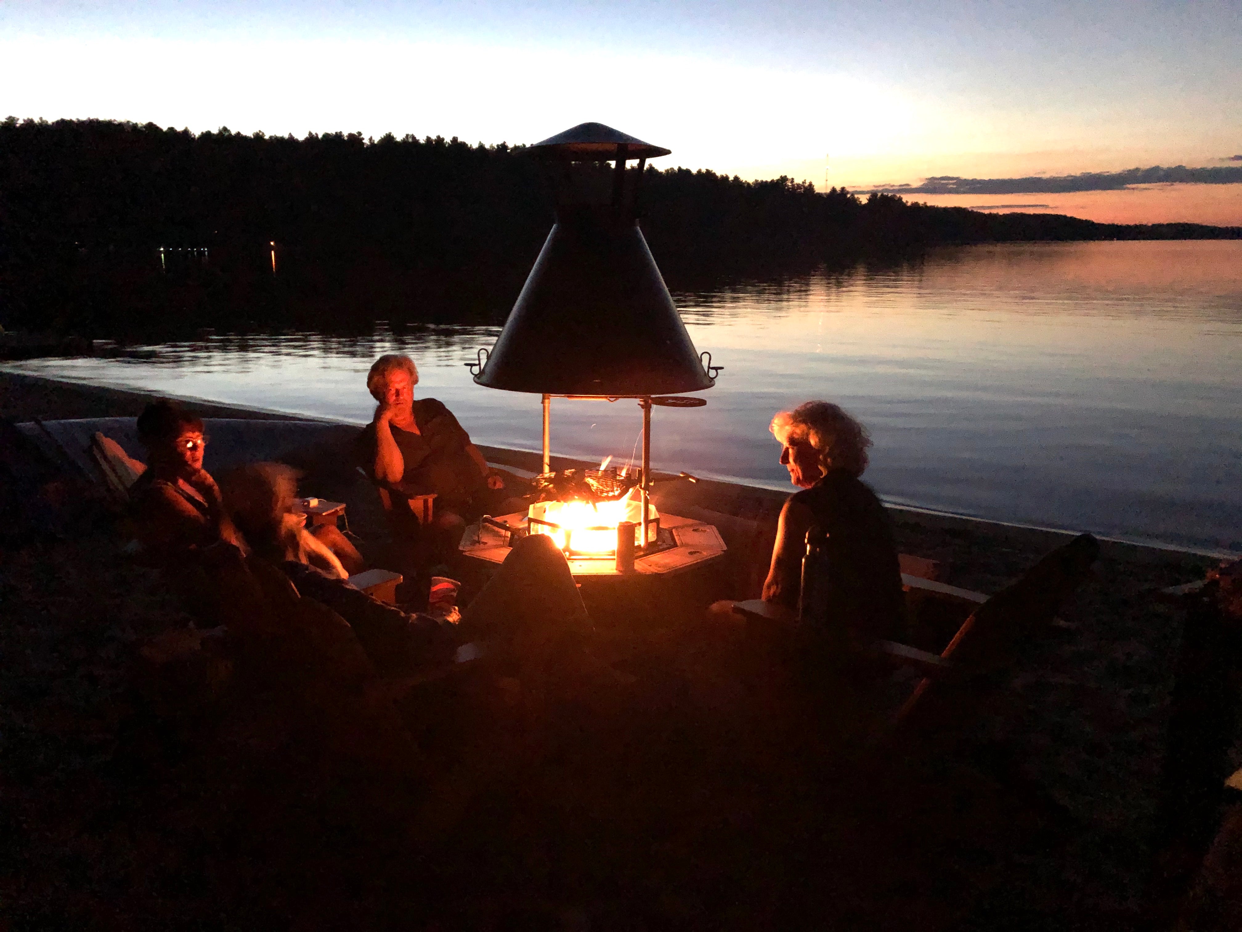       A group of people sit around a Kota Grill on a beach at dusk. 